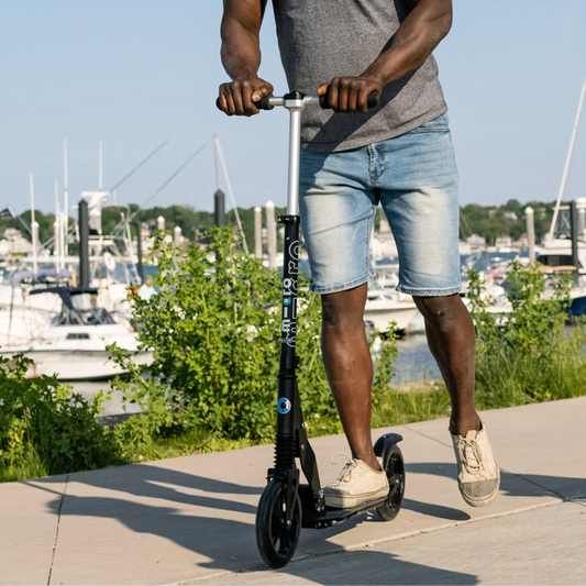 A man riding a black electric scooter on a paved path outdoors near a marina, wearing a gray henley shirt, denim shorts, and sneakers.