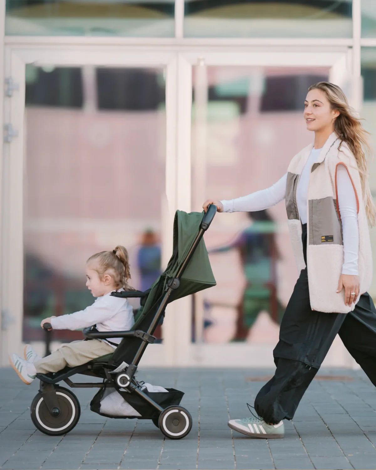 Woman pushing a green Smartrike Traveler with a child on a sidewalk.