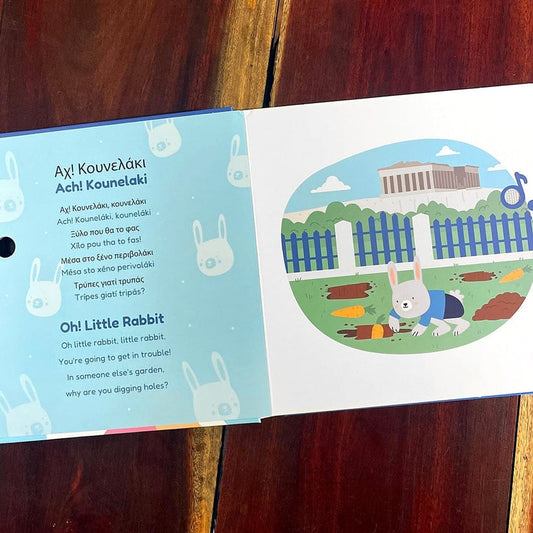 An open children's book featuring the Greek nursery rhyme *"Ach! Kounselaki"* (*"Oh! Little Rabbit"*), showing song lyrics alongside an illustration of a rabbit digging in a garden with the Parthenon in the background.