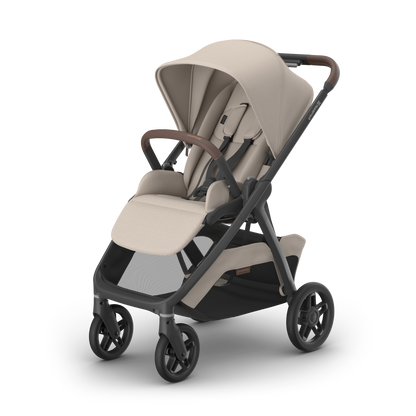 A modern stroller with a light beige fabric canopy and seat, a dark gray frame, and brown leather-look handlebar grips, shown on a clean white background.