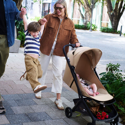 A family of four walks along a tree-lined sidewalk. A woman in a brown jacket and white pants pushes a beige stroller with a baby inside, while a man in a blue shirt holds a young boy in a striped shirt and khaki pants.
