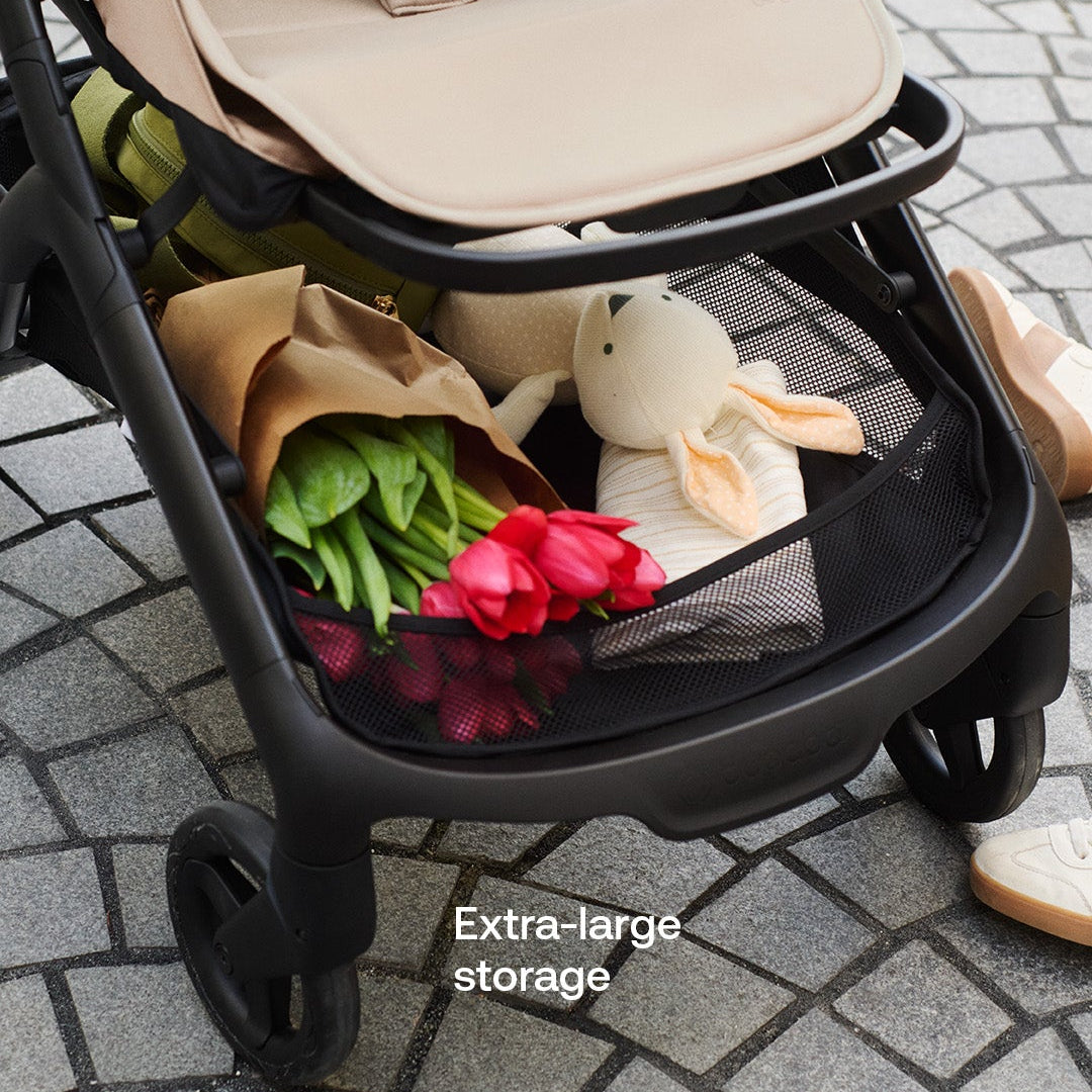 A beige and black baby stroller on a gray cobblestone street, showing its extra-large storage basket filled with flowers, a plush toy, and a shoe, with the text 'Extra-large storage' overlaid.