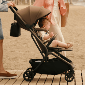 A baby stroller with a child sitting inside, being pushed by an adult on a wooden boardwalk at the beach. The stroller is brown and black.