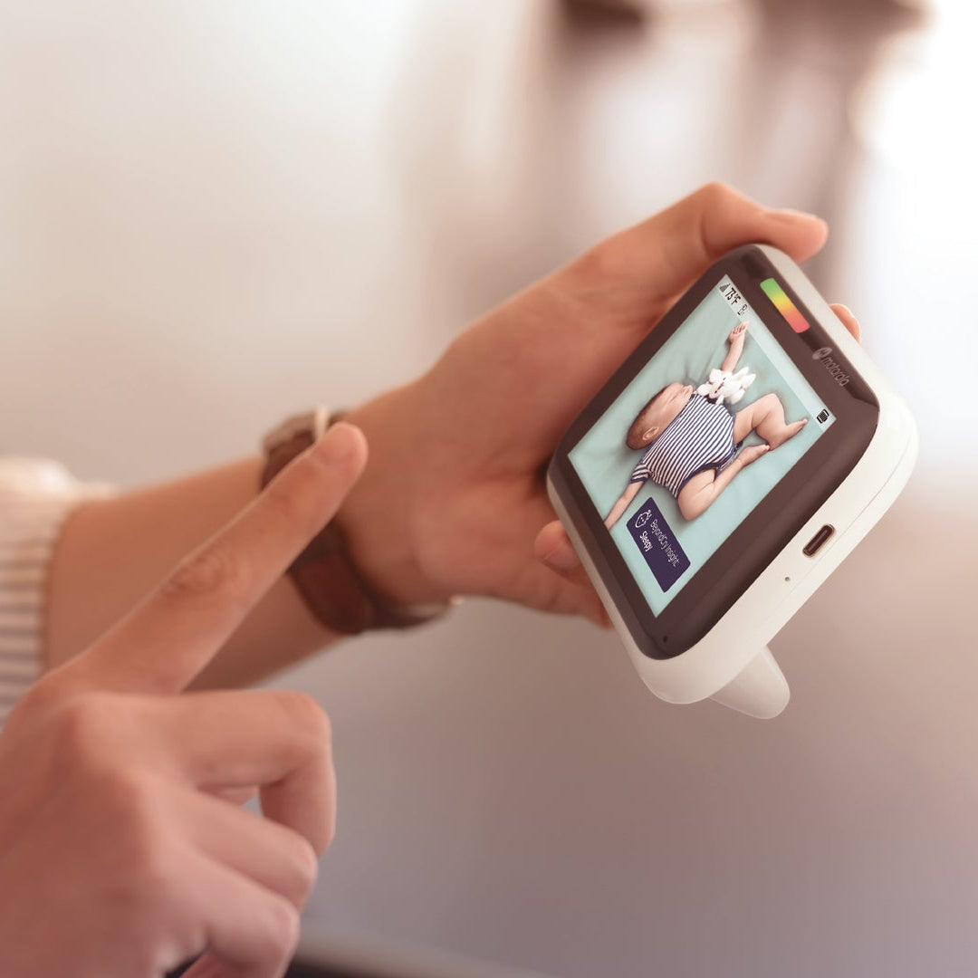 A close-up of a person holding a handheld baby monitor with a small screen showing a baby, with one hand pointing at the display. The person is wearing a cream-colored ribbed long-sleeve shirt, and the background is a softly blurred indoor setting.