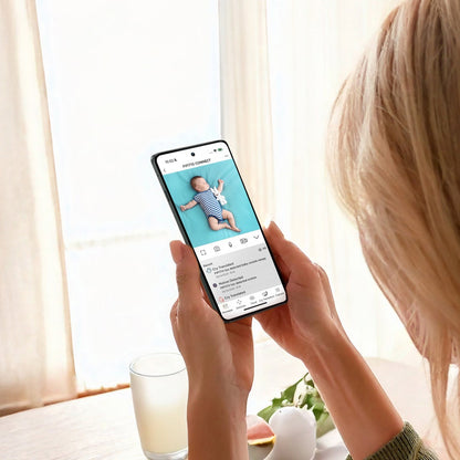 A woman with blonde hair holds a smartphone showing a baby care or sleep app screen, photographed from behind in a bright indoor setting near a window with curtains.