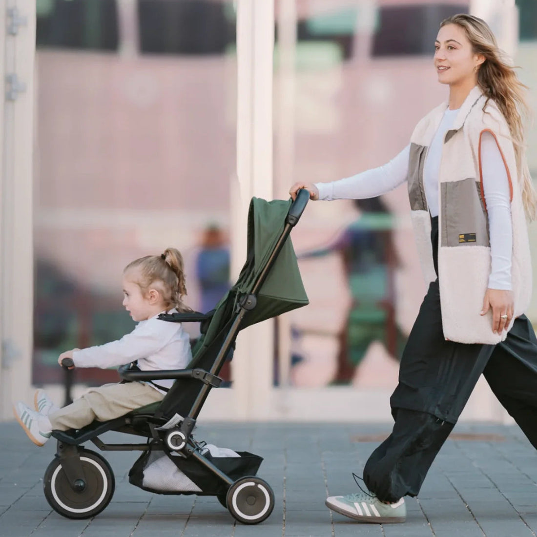 Woman pushing a green Smartrike Traveler with a child on a sidewalk.