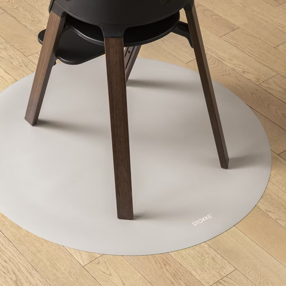 
A round off-white protective floor mat under a black chair with dark wood legs, placed on a light-colored wooden floor. The brand name 'STOCKE' is printed in small white letters on the bottom right edge of the mat.