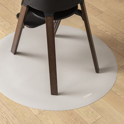 
A round off-white protective floor mat under a black chair with dark wood legs, placed on a light-colored wooden floor. The brand name 'STOCKE' is printed in small white letters on the bottom right edge of the mat.