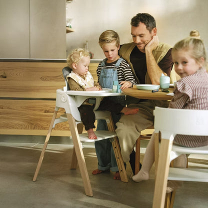 
A family scene in a modern kitchen with a man seated at a wooden table with two children in white high chairs, one on each side. The setting features warm wood cabinetry and a light gray floor.
