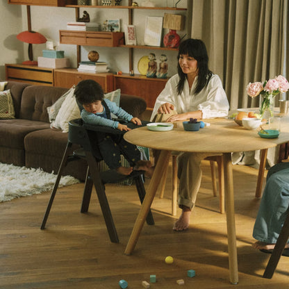 
A woman and a child are seated in a warm, modern living room. The child is sitting in a black high chair next to a round wooden dining table, while the woman sits on a wooden chair across from the table.