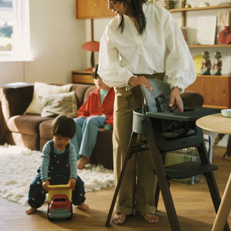 
A woman in a white blouse and khaki pants stands barefoot in a living room next to a dark gray high chair, while a young child plays with a toy truck on the wooden floor in the foreground.