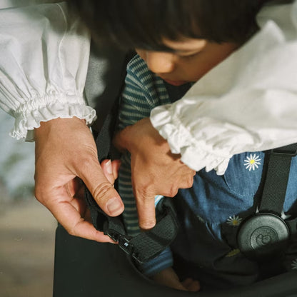 
A close-up lifestyle shot of a child wearing a blue denim bib with a white floral print, seated in a dark carrier or seat, while an adult's hands fastens the black safety harness strap. The child wears a striped shirt underneath and a round black ID tag is attached to the harness.