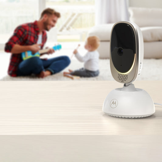 A Motorola baby monitor camera with a white base and a silver-and-black oval face sits on a light wood table in the foreground, with a blurred living room scene behind it showing a man and child playing.