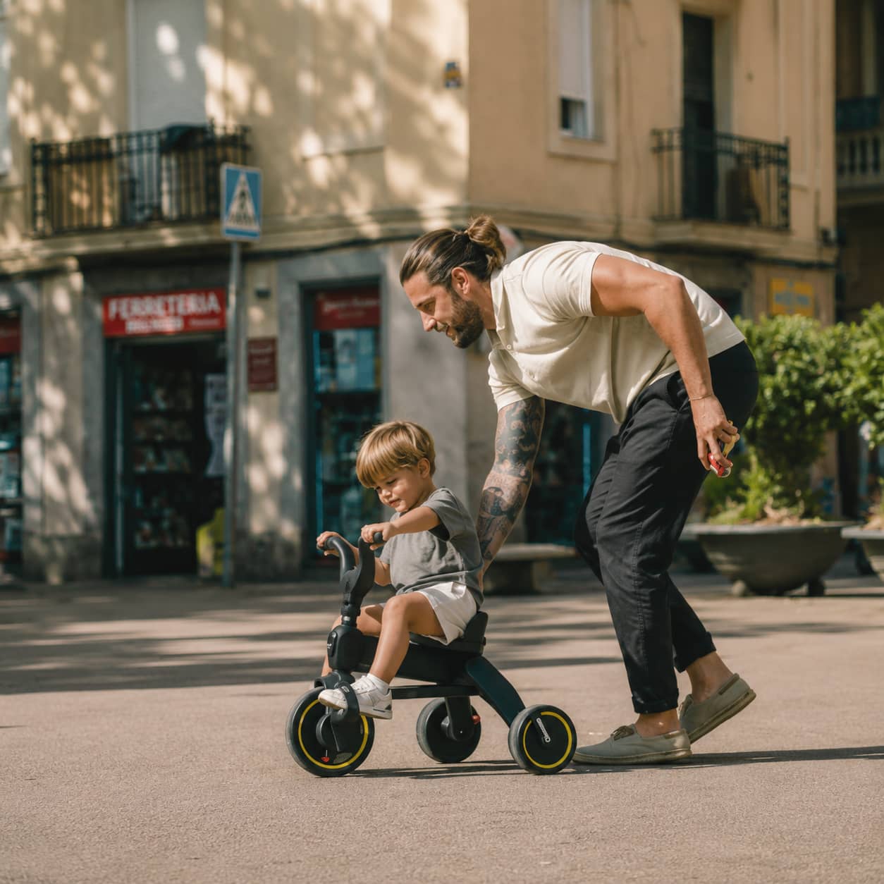 A father helping his young son ride a black Doona trike on a sunny city street.