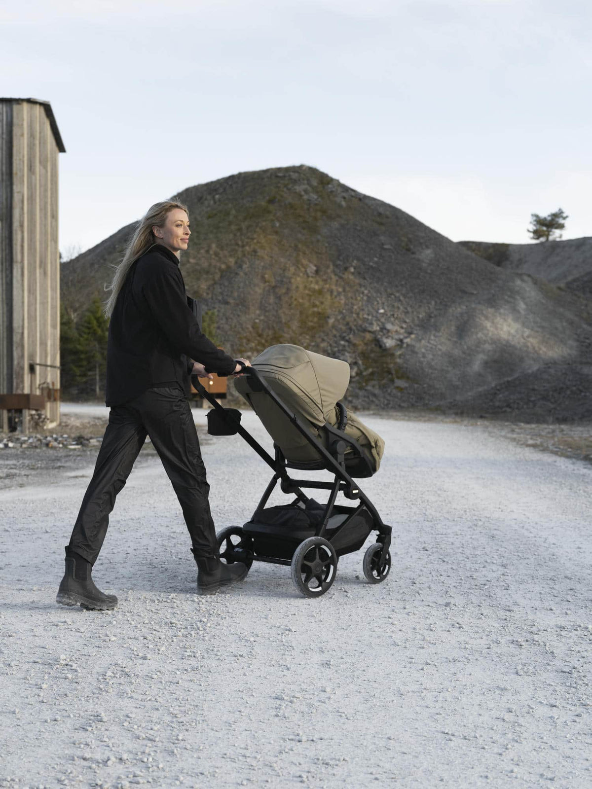 Woman pushing a khaki Thule Charm stroller in toddler-seat mode outdoors.
