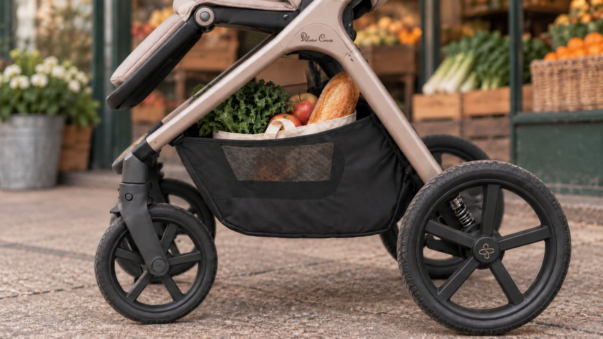A close-up of a stroller with a beige frame and black wheels, featuring a black mesh side storage basket filled with groceries like vegetables and bread. The stroller is positioned outdoors on a paved surface with a blurred market or garden store background.