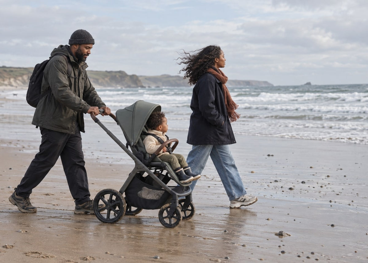 A man pushes a stroller with a child on a beach while a woman walks beside them. The stroller has a dark gray canopy and frame with black wheels, and the scene is set on a sandy shoreline with the ocean in the background.