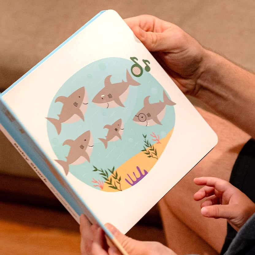
A person holding open a children's board book with shark illustrations while a young child reaches toward the pages.