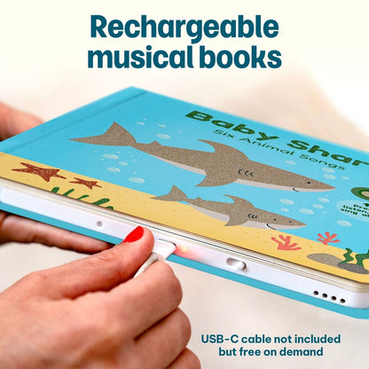 
A close-up of a person holding a colorful children's book with a shark underwater illustration and text reading 'Baby Shark' and 'Six Animal Songs.' A USB-C cable is being plugged into the book, and text at the top says 'Rechargeable musical books' and text at the bottom says 'USB-C cable not included but free on demand.