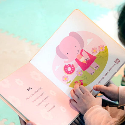 
Child's hand holding a book with a colorful illustration of a girl and a donut on a pink background.