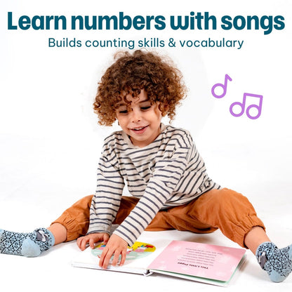 
A young child with curly brown hair sits on a white floor, reading a pink book while wearing a gray and white striped long-sleeve shirt, rust-orange pants, and blue patterned socks. The image includes promotional text at the top reading 'Learn numbers with songs' and 'Builds counting skills & vocabulary,' plus purple music note icons.