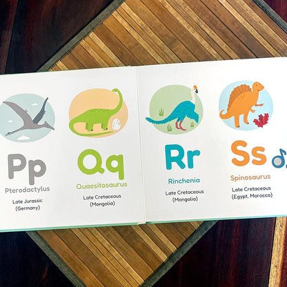 
An open children's educational book showing the letters P, Q, R, and S with dinosaur illustrations and names, placed on a striped wooden surface.