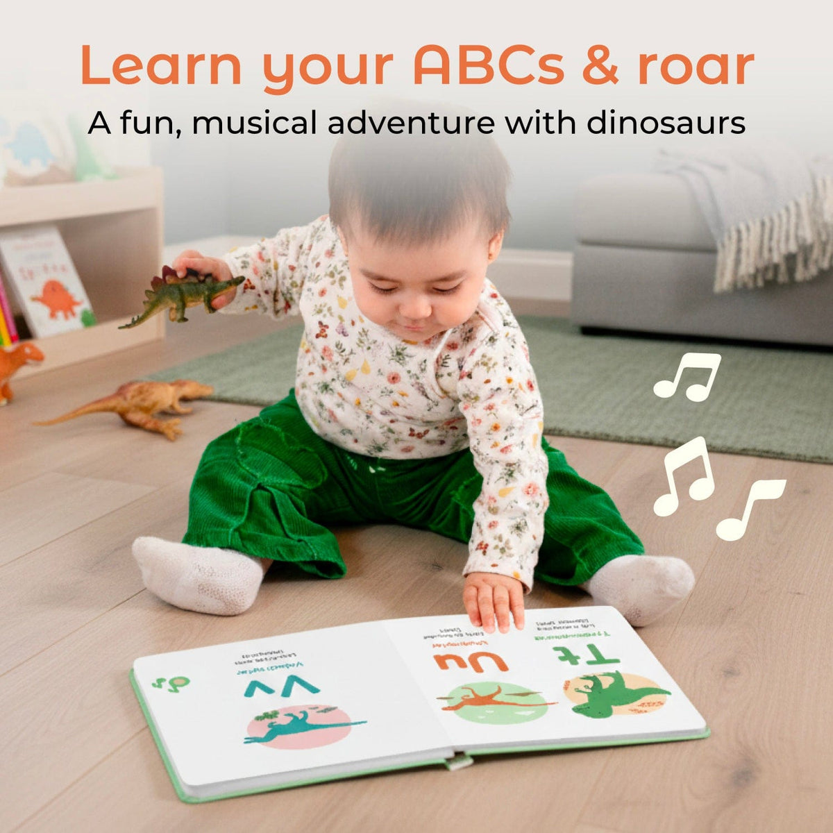 
A baby sitting on a wooden floor in a playroom, reading a colorful interactive children's book about dinosaurs. The baby is wearing a cream floral long-sleeve top and green pants.