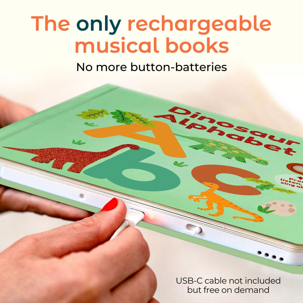 
A person holds a light green children's book titled 'Dinosaur Alphabet' while plugging a USB-C cable into it. The cover features colorful dinosaur illustrations and large letters A, B, and C.
