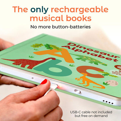 
A person holds a light green children's book titled 'Dinosaur Alphabet' while plugging a USB-C cable into it. The cover features colorful dinosaur illustrations and large letters A, B, and C.