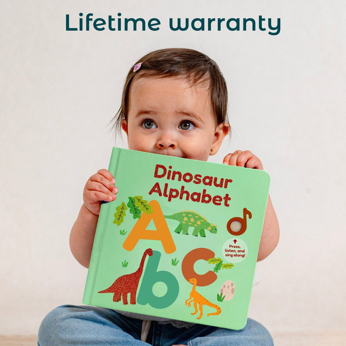 
A baby girl holds a green 'Dinosaur Alphabet' book in front of her face, with the text 'Lifetime warranty' written at the top. The book features colorful dinosaur illustrations and letters.