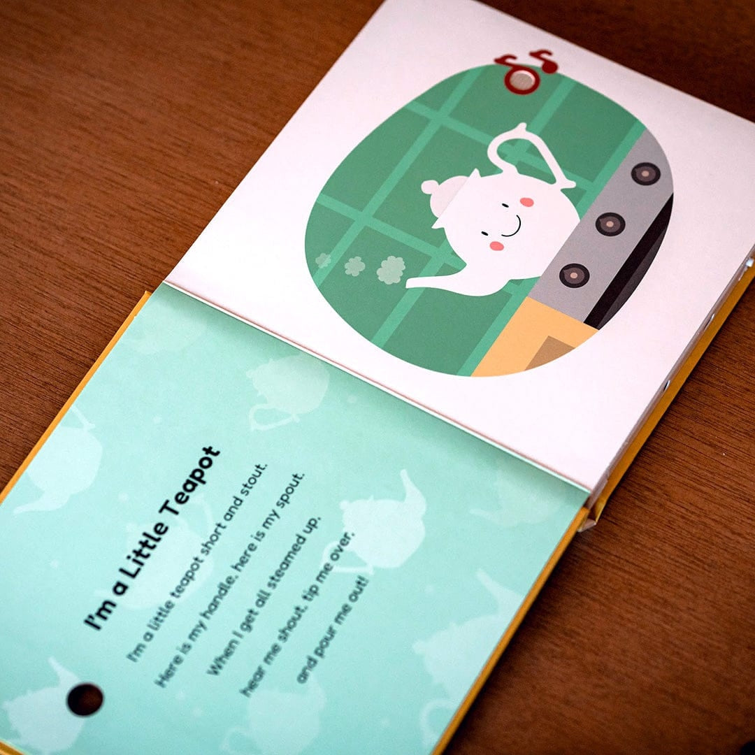 
A children's book or card set photographed on a wooden surface, showing a pastel illustration of a teapot and the text 'I'm a Little Teapot' on the lower page.
