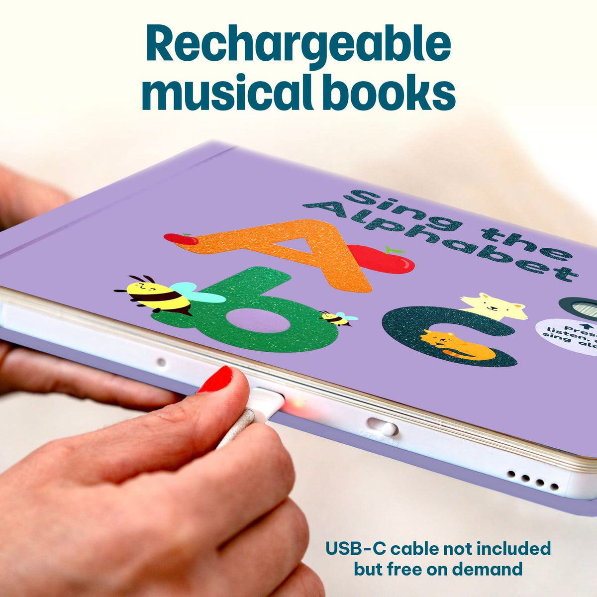 
A close-up of a hand plugging a USB-C cable into a lavender educational children's book titled 'Sing the Alphabet' with colorful alphabet letters and animal illustrations. Text overlays read 'Rechargeable musical books' and 'USB-C cable not included but free on demand'.