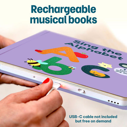 
A close-up of a hand plugging a USB-C cable into a lavender educational children's book titled 'Sing the Alphabet' with colorful alphabet letters and animal illustrations. Text overlays read 'Rechargeable musical books' and 'USB-C cable not included but free on demand'.