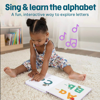 
A toddler sits on a cream shag rug reading an interactive alphabet book. The scene includes promotional text at the top reading 'Sing & learn the alphabet' and purple musical note graphics.