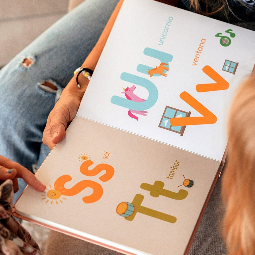 
A close-up of a children's alphabet book being held by a person wearing ripped blue jeans. The open pages show colorful uppercase letters with playful illustrations and Spanish words, including 'unicomio', 'ventana', 'sol', and 'tambor'.