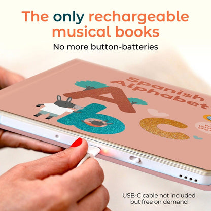 
A close-up of hands charging a pink Spanish Alphabet musical book with a USB-C cable. The book cover features colorful glittery letters and cartoon animals.