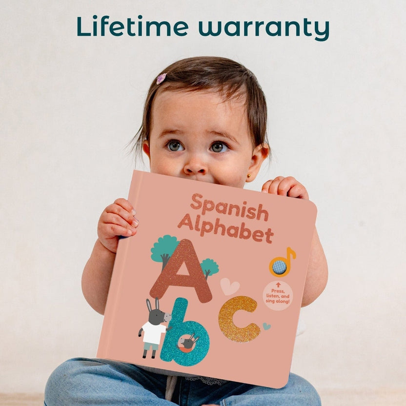 
A baby sits holding a Spanish Alphabet book, with the words 'Lifetime warranty' printed at the top of the image. The book has a dusty pink cover decorated with colorful illustrations of the Spanish alphabet letters.