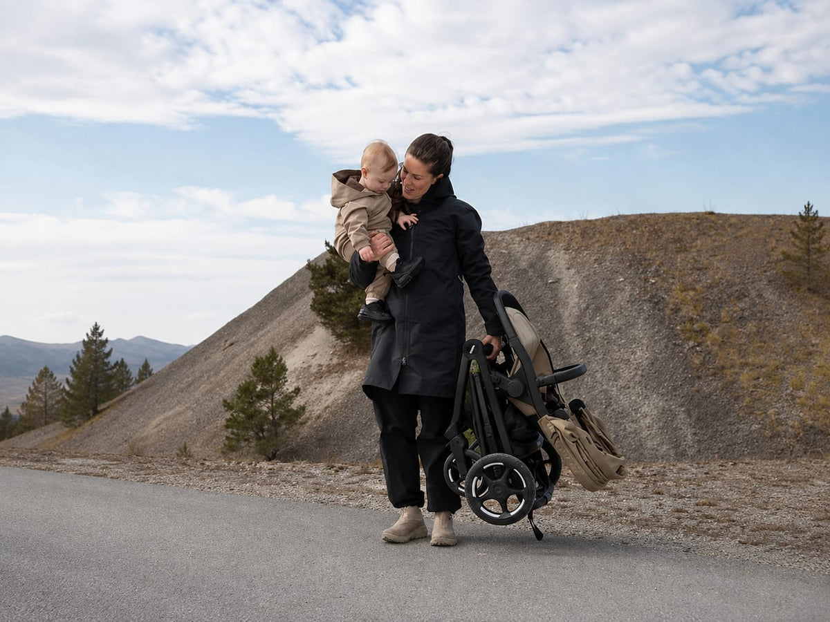 Mother carrying a toddler while easily carrying the compactly folded Thule Charm stroller in one hand.
