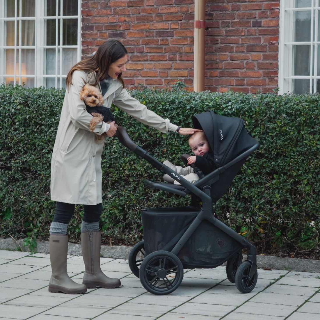Woman walking outdoors with a baby in a Nuna DEMI Icon stroller and a small dog in her arms.
